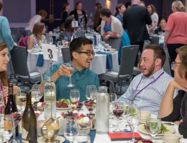 Two men engaged in conversation around a dinner table with wine, salads and dessert already set. Two other guests lean into their conversations. The man wearing a green shirt has a glass of wine in his hand.
