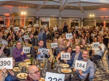 Room full of happy and excited guests sitting around dinner tables while eating and raising bid paddles