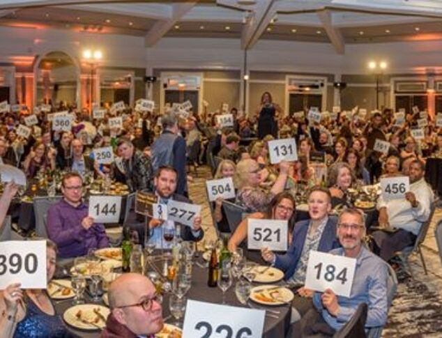 Room full of happy and excited guests sitting around dinner tables while eating and raising bid paddles