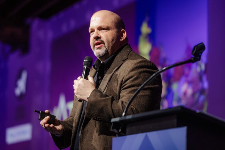 T. Clay Buck speaking on stage in front of a podium while holding a microphone against a purple background at the Elevate 2026 conference on fundraising events