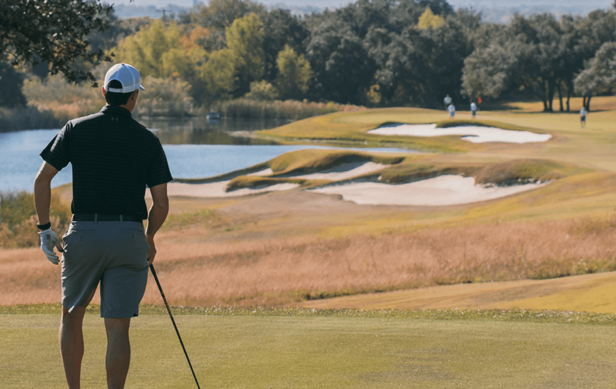 A man in golf attire holding a golf club stands on a golf course looking out a fairway with long grass on the edges, sand bunkers and a small lake