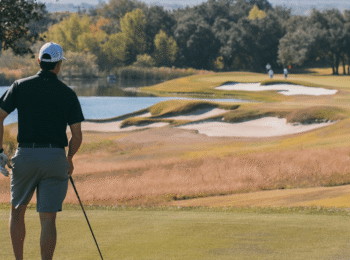 A man in golf attire holding a golf club stands on a golf course looking out a fairway with long grass on the edges, sand bunkers and a small lake