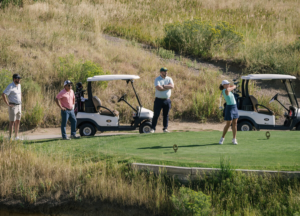 Four players on a golf course with a female player in mid-swing and a two golf carts in the background