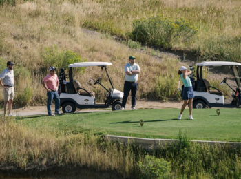 Four players on a golf course with a female player in mid-swing and a two golf carts in the background